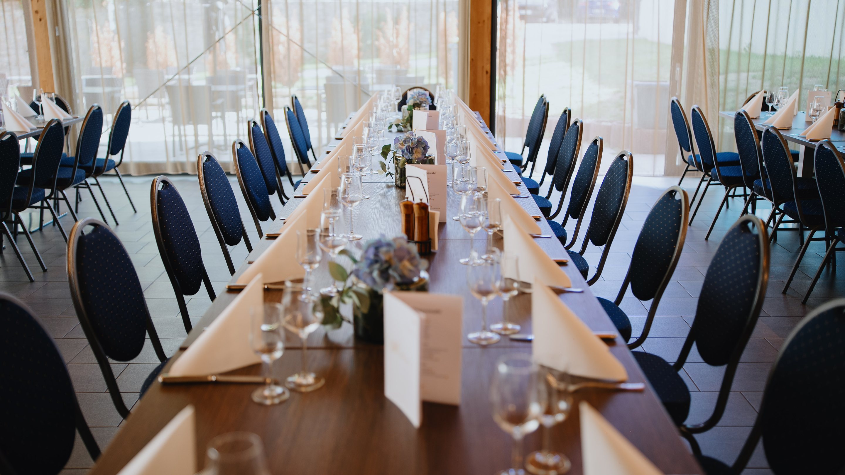Lange, festlich eingedeckte Tafel mit Bankettstühlen vor einer Glasfront